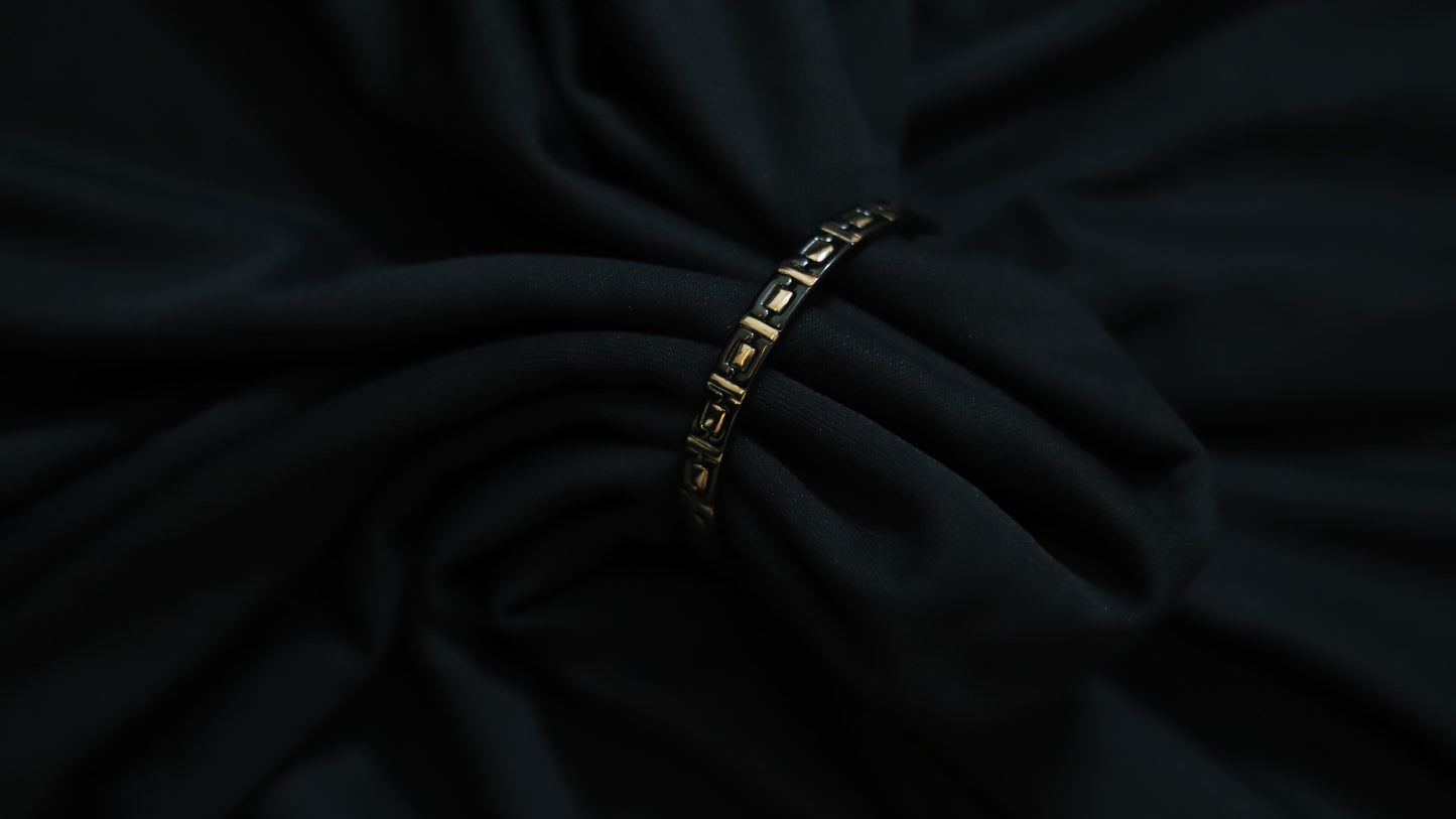 A close-up of a black fabric garment with a thin gold chain bracelet threaded through the center, set against a dark draped background.