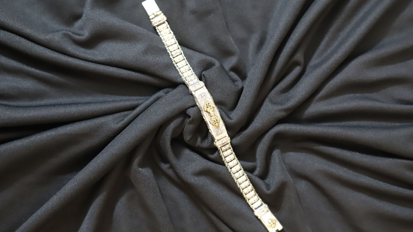 A silver-toned metal link bracelet with a central gold-toned decorative emblem, laid diagonally over draped black fabric.