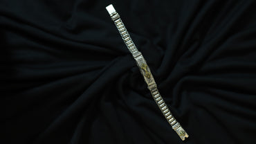 A silver-toned metal bracelet with textured rectangular links and gold-toned decorative details, displayed diagonally on a draped black fabric background.