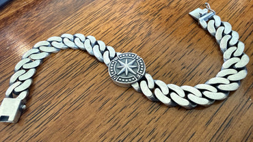 A silver-toned Cuban link chain bracelet with a central circular medallion featuring a starburst design, displayed on a wooden surface.
