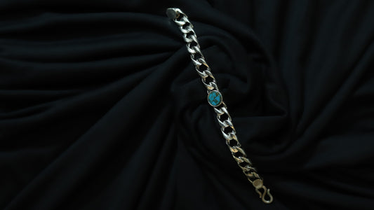 A silver-toned curb chain bracelet with a small round turquoise blue stone accent, displayed on a draped black fabric background.