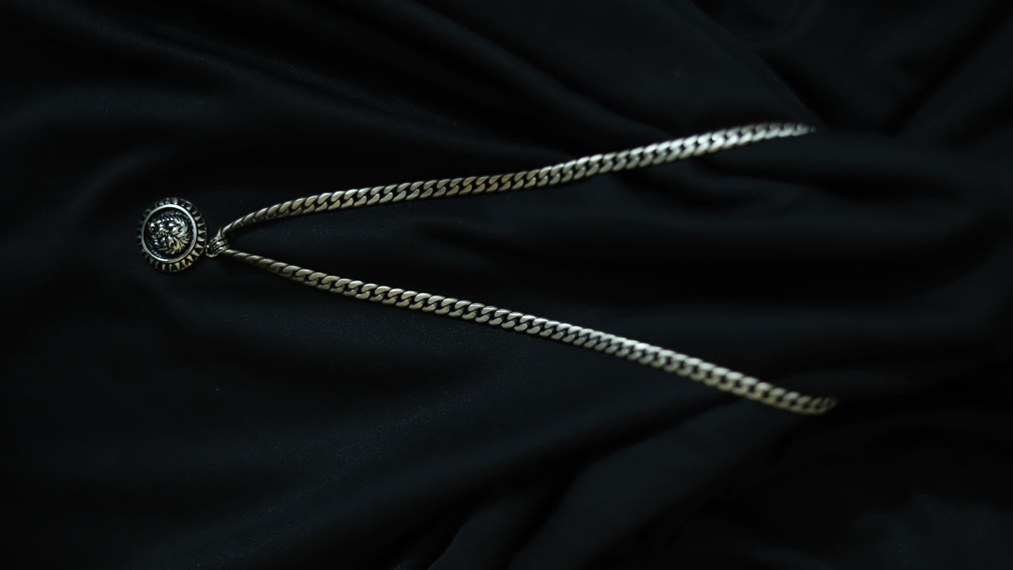 A silver-toned chain necklace with a circular medallion pendant featuring a stylized face, laid out on a dark black fabric background.