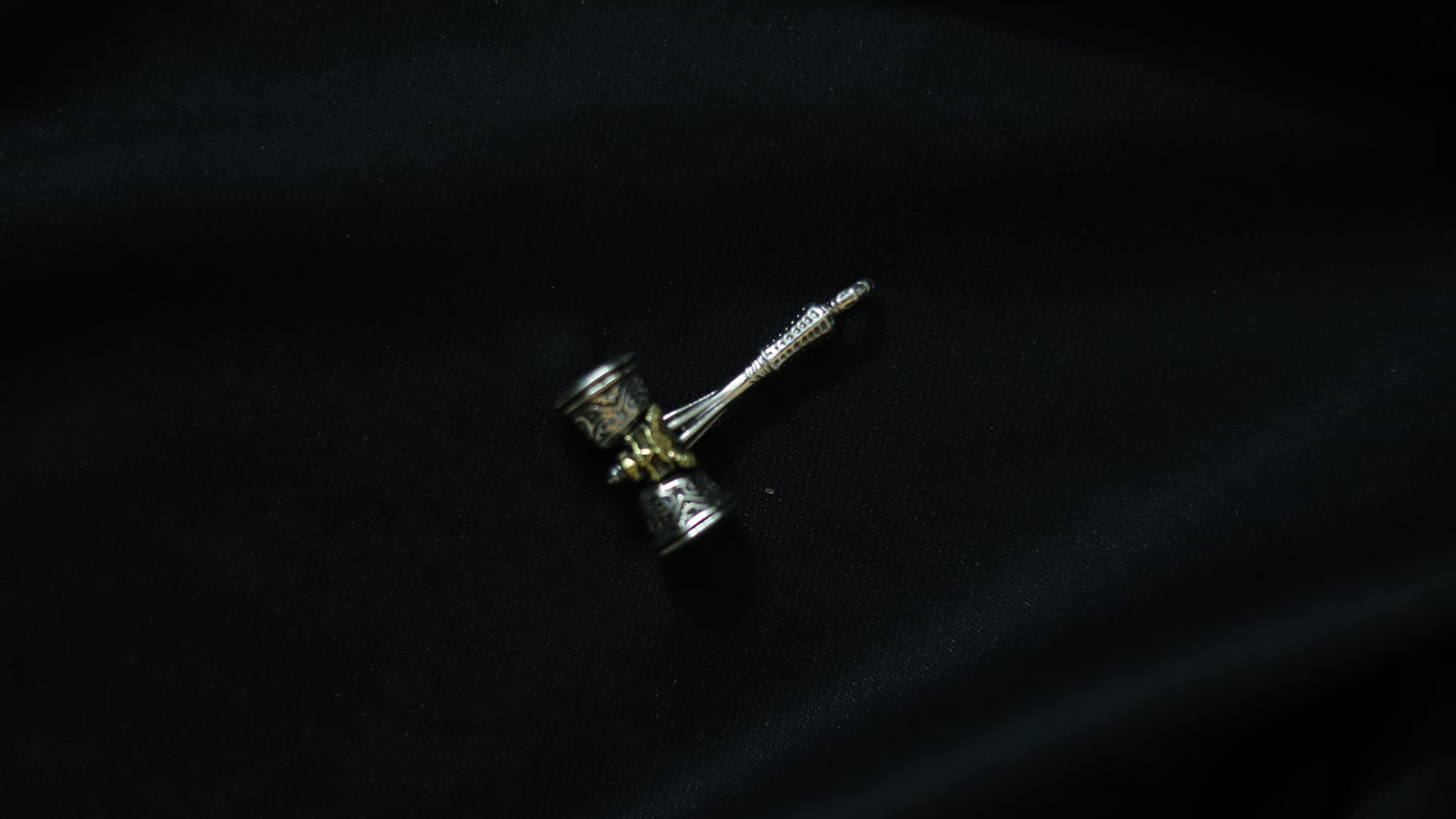 A miniature hammer-shaped accessory with a silver-toned handle and head, featuring gold-toned detailing, resting on a dark black fabric background.