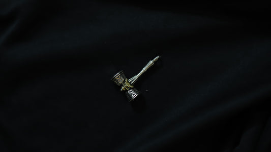 A small silver-toned metal pendant or charm with a textured cylindrical head and a long pointed stem, photographed on a dark black fabric background.