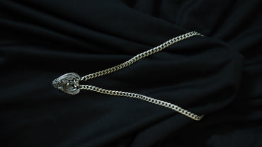 A silver-toned chain necklace with a decorative, textured pendant, laid out on a draped black fabric background.
