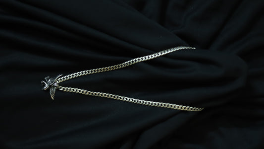 A silver-toned metal chain necklace with a small winged bird pendant, laid out on a draped black fabric background.