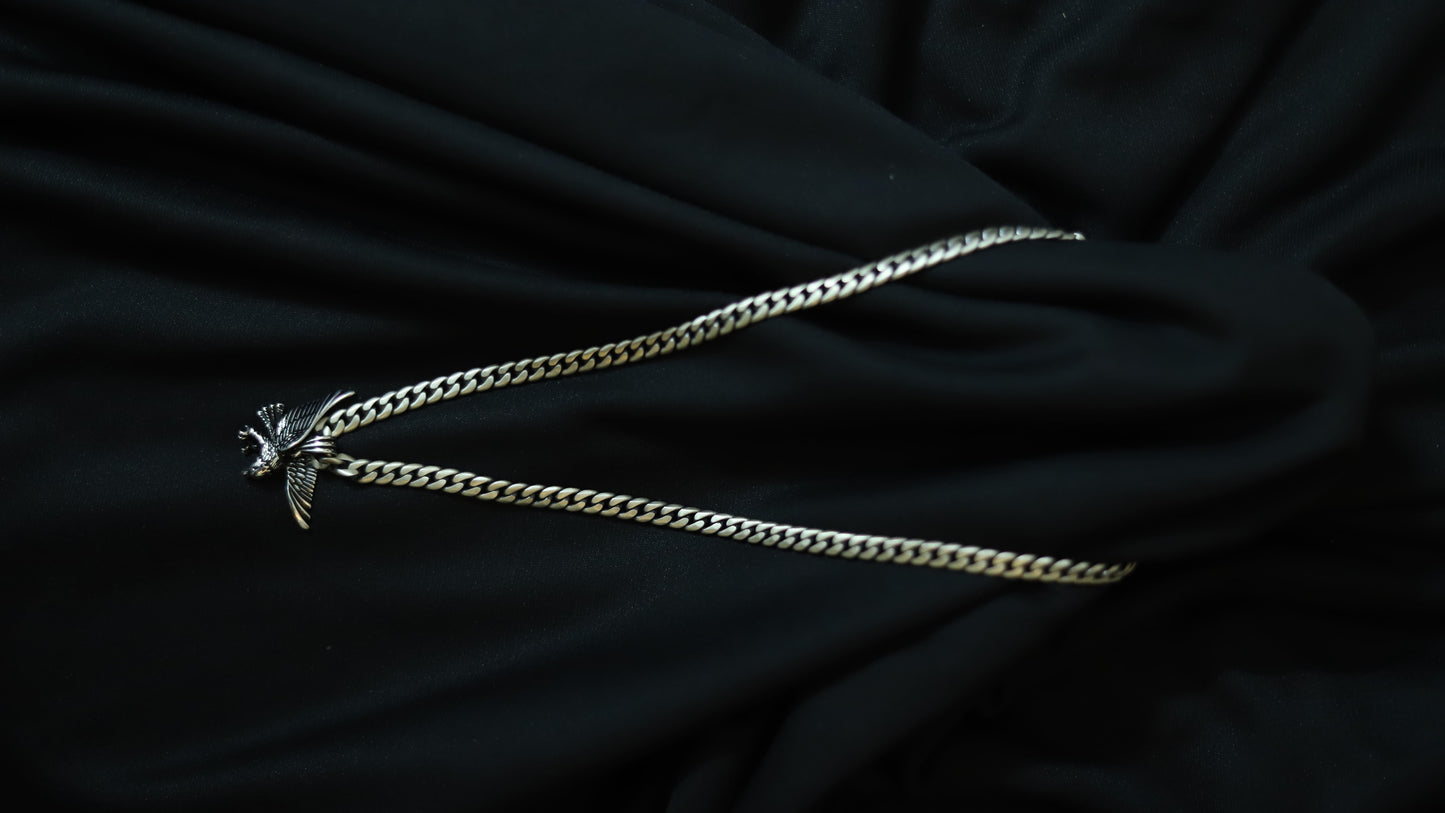 A silver-toned curb chain necklace with a small bird pendant, laid out on draped black fabric.