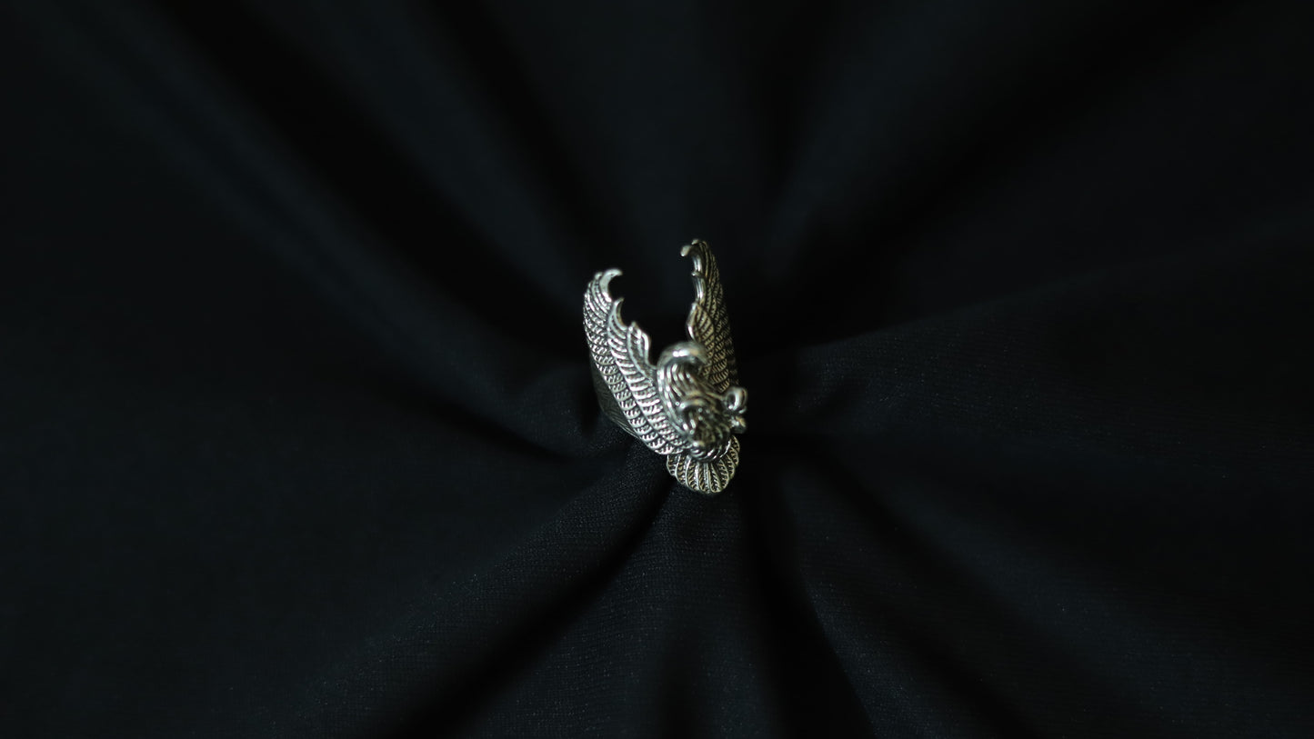 A silver-toned metal ring shaped like a stylized eagle or bird head with wings, centered on a dark black fabric background.