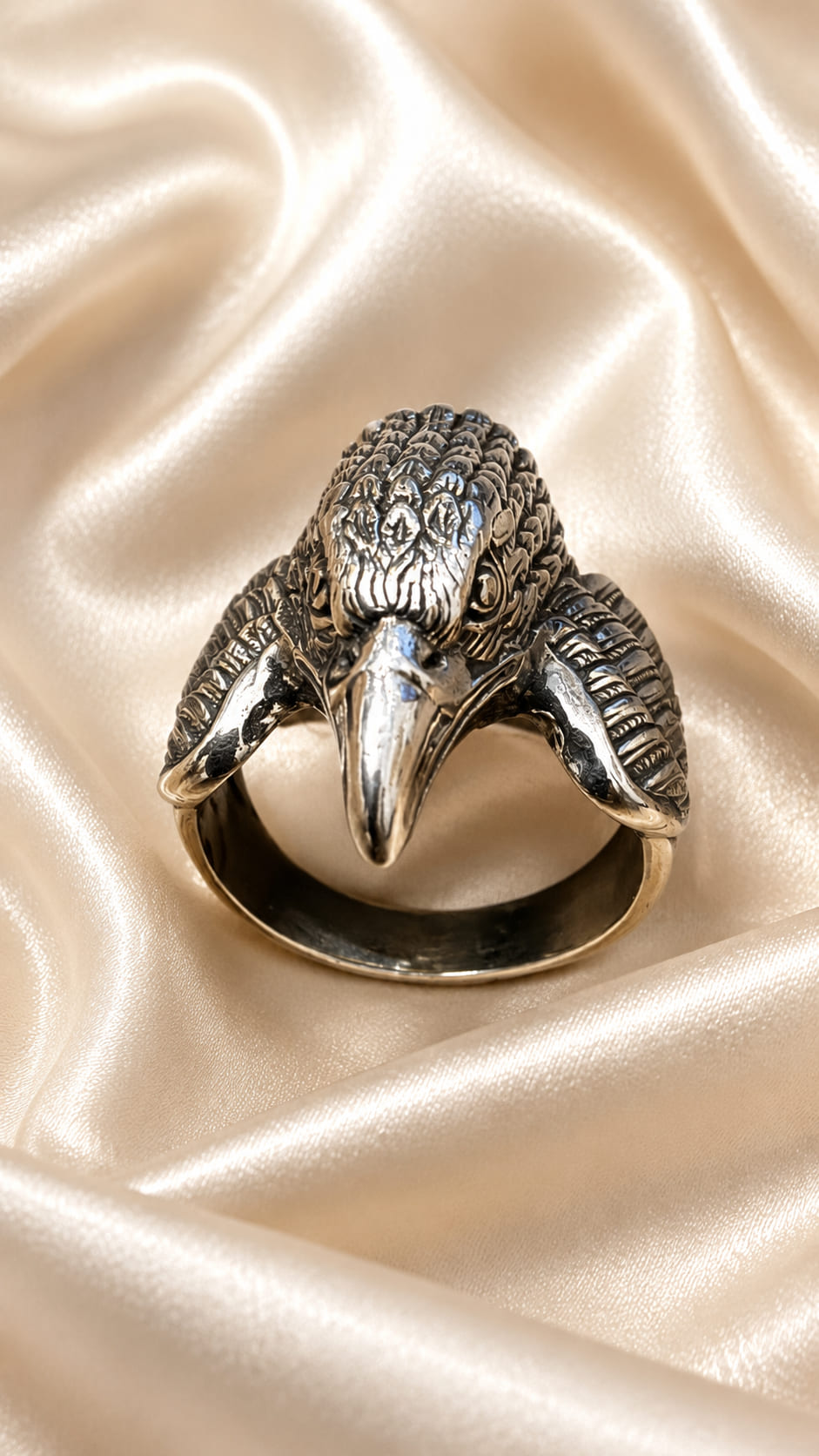A silver-toned ring shaped like a raven or eagle head, displayed on draped champagne satin fabric.