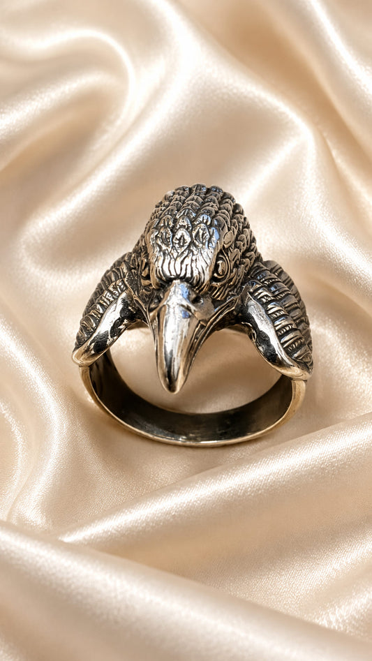 A silver-toned ring shaped like a raven or eagle head, displayed on draped champagne satin fabric.