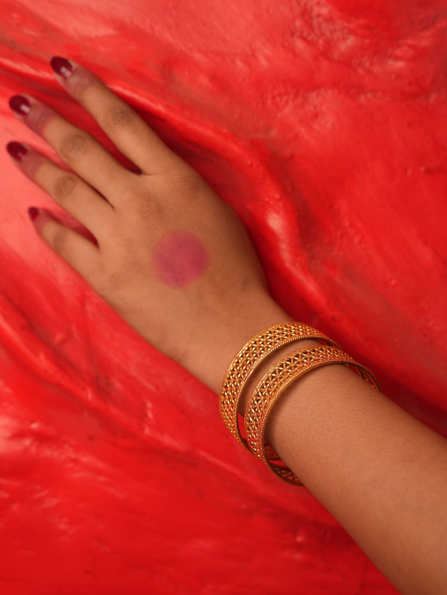 A close-up of a person's hand resting on a textured red surface, wearing three gold bangles (kara bracelets) and having dark red nail polish.