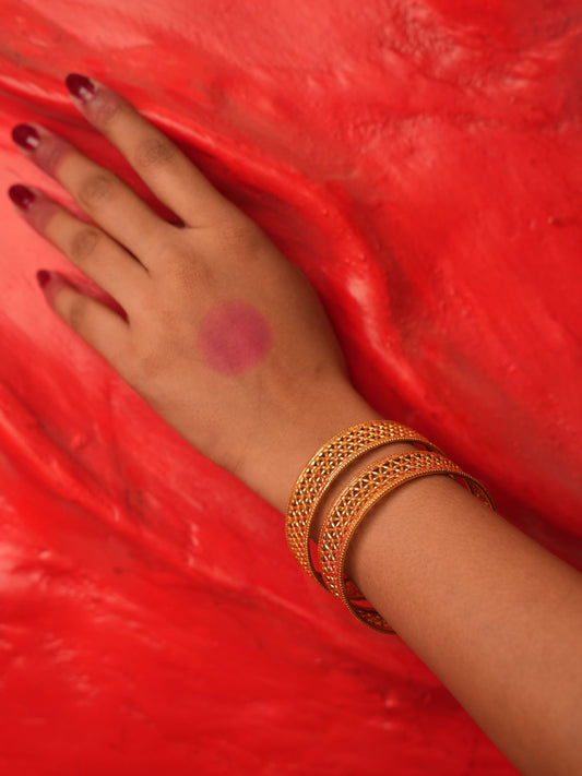 A close-up of a person's hand resting on a textured red surface, wearing three gold bangles (kara bracelets) and having dark red nail polish.