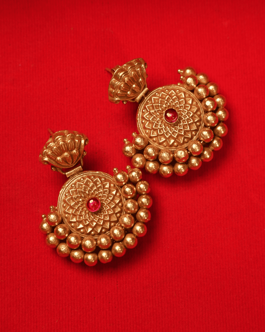 A pair of traditional Indian gold-toned earrings with red stone accents, displayed on a textured red background.