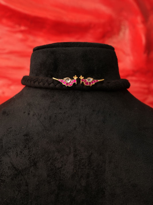 A black braided hair accessory with two gold-toned bird charms embellished with pink and green enamel, displayed on a black velvet mannequin bust against a red fabric background.