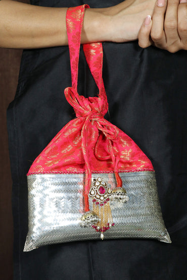 A small drawstring pouch bag with a red and gold patterned top half and a silver woven bottom half, decorated with a decorative tassel charm, held against a black garment by a person's hands and arm.