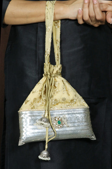 A close-up of a person holding a small drawstring pouch bag. The bag has a beige and gold patterned upper half, a metallic silver patterned lower half, and a decorative silver clasp with a green stone.