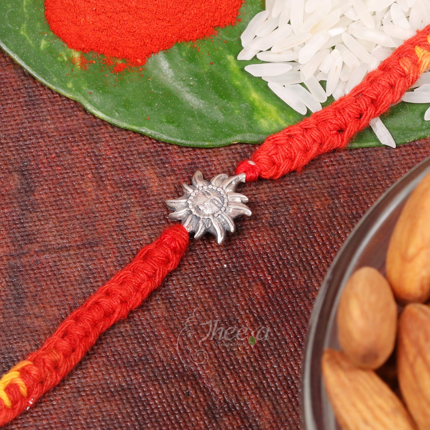 A red braided Rakhi bracelet with a silver sun-shaped charm, displayed on a textured dark brown surface with festive props including a green leaf, red powder, white rice, and almonds.