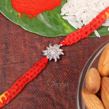A red braided Rakhi bracelet with a silver sun-shaped charm, displayed on a textured dark brown surface with festive props including a green leaf, red powder, white rice, and almonds.