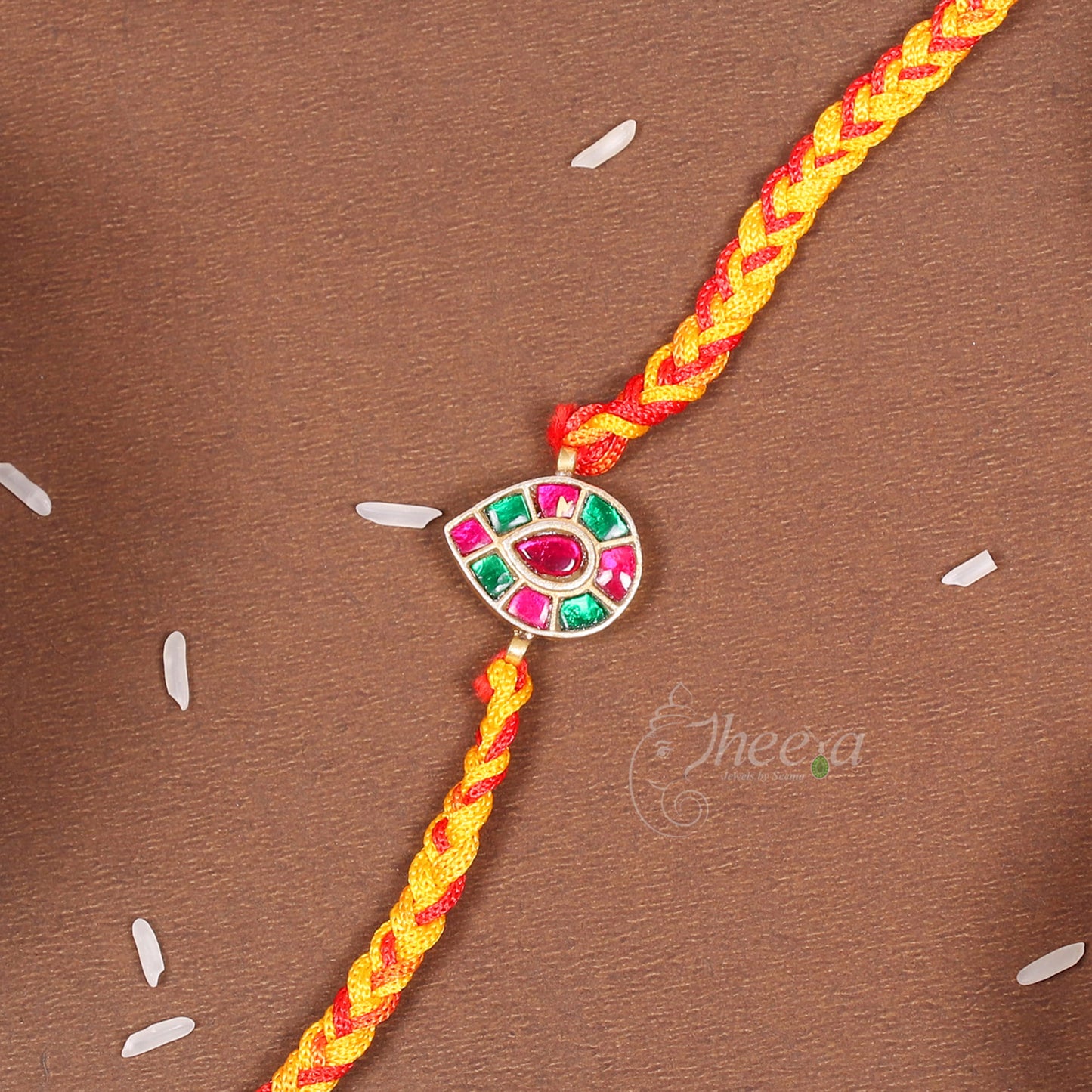 A braided yellow and red cord Rakhi bracelet with a central silver-toned evil eye charm embellished with pink and green stones, laid diagonally on a brown textured background.