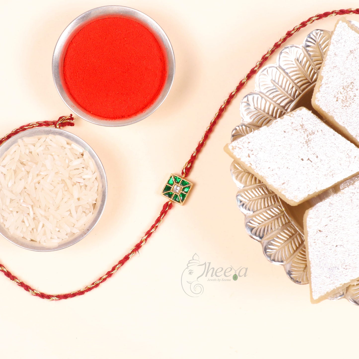 A traditional Indian Rakhi bracelet with a red and gold braided cord and a central green and gold square charm, displayed alongside rice, red powder, and sweets on a light beige background.