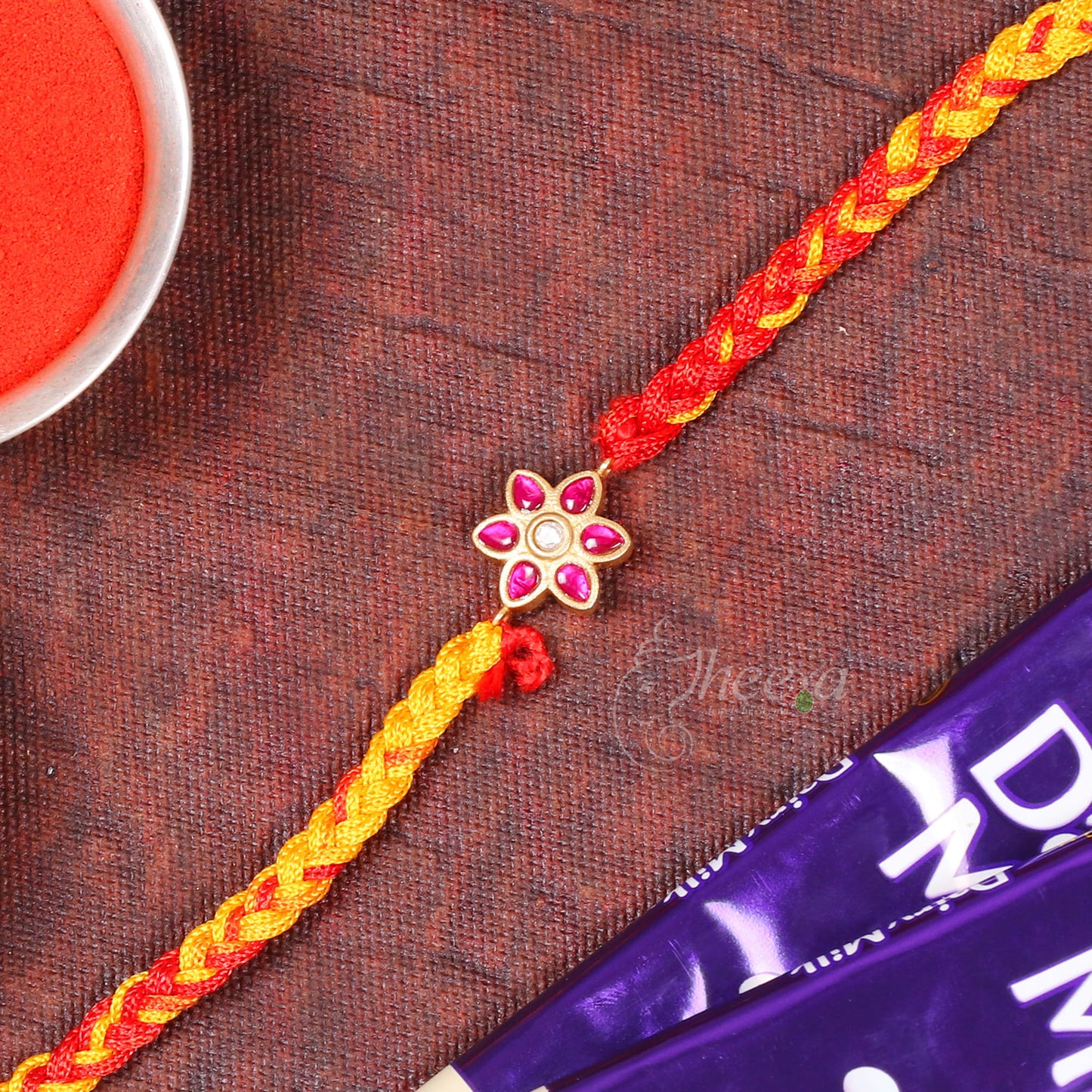 A braided Rakhi bracelet with a gold-tone flower charm set with pink stones, laid diagonally on a textured dark brown surface with a red powder dish and chocolate bar partially visible.