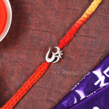A braided Rakhi featuring a red and yellow gradient cord with a silver Om symbol charm, laid diagonally on a textured brown fabric background with a bowl of red powder and a purple packet partially visible.
