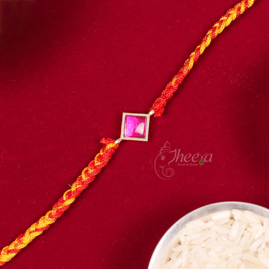 A red and yellow braided Rakhi bracelet with a central square pink stone in a gold frame, displayed diagonally on a dark red background with a bowl of rice partially visible in the corner.