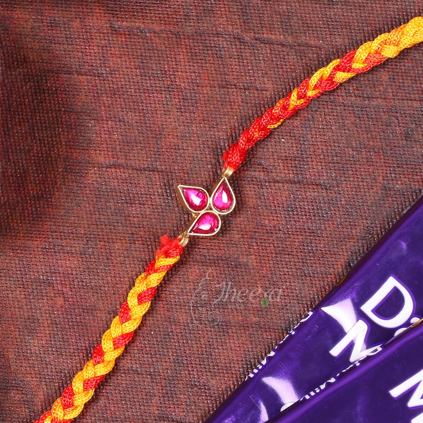 A braided Rakhi bracelet with red and yellow threads and a gold-toned butterfly charm with pink stones, laid diagonally on a textured brown fabric next to a purple chocolate bar.