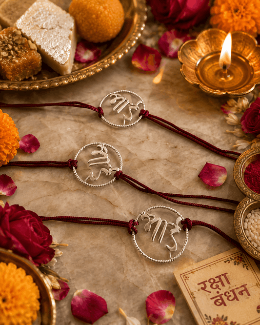 Three silver wire Rakhi bracelets with Devanagari script are arranged on a beige stone surface with marigold flowers, rose petals, a lit diya, and traditional sweets. A card reading 'Rakhi Bandhan' is visible in the lower right corner.