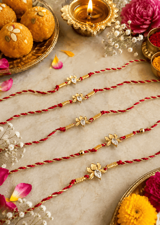 Four red and gold thread Rakhi bracelets are arranged diagonally on a beige marble surface, surrounded by festive props including laddoo, a lit diya, and flowers.
