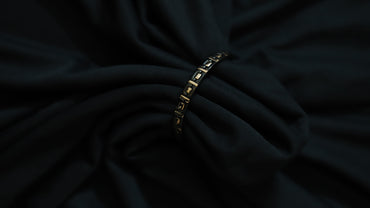 A close-up of a black fabric garment with a thin gold chain bracelet threaded through the center, set against a dark draped background.