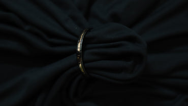 A slim metallic bangle bracelet with a geometric pattern, shown tied around a dark, draped fabric background.