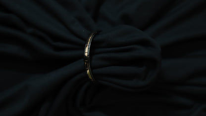 A slim metallic bangle bracelet with a geometric pattern, shown tied around a dark, draped fabric background.