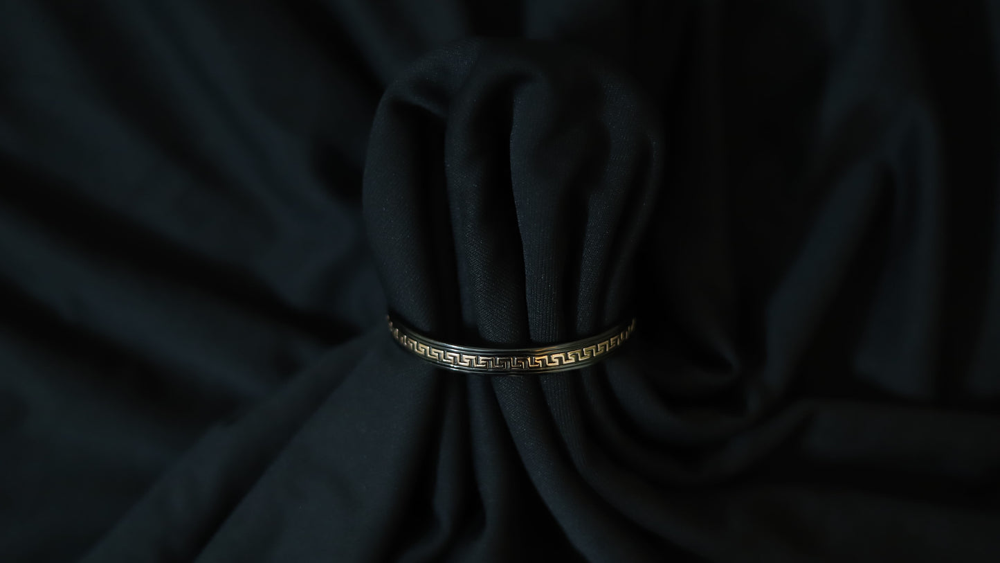A black bangle bracelet with a gold Greek key pattern, wrapped around a dark black fabric bundle.