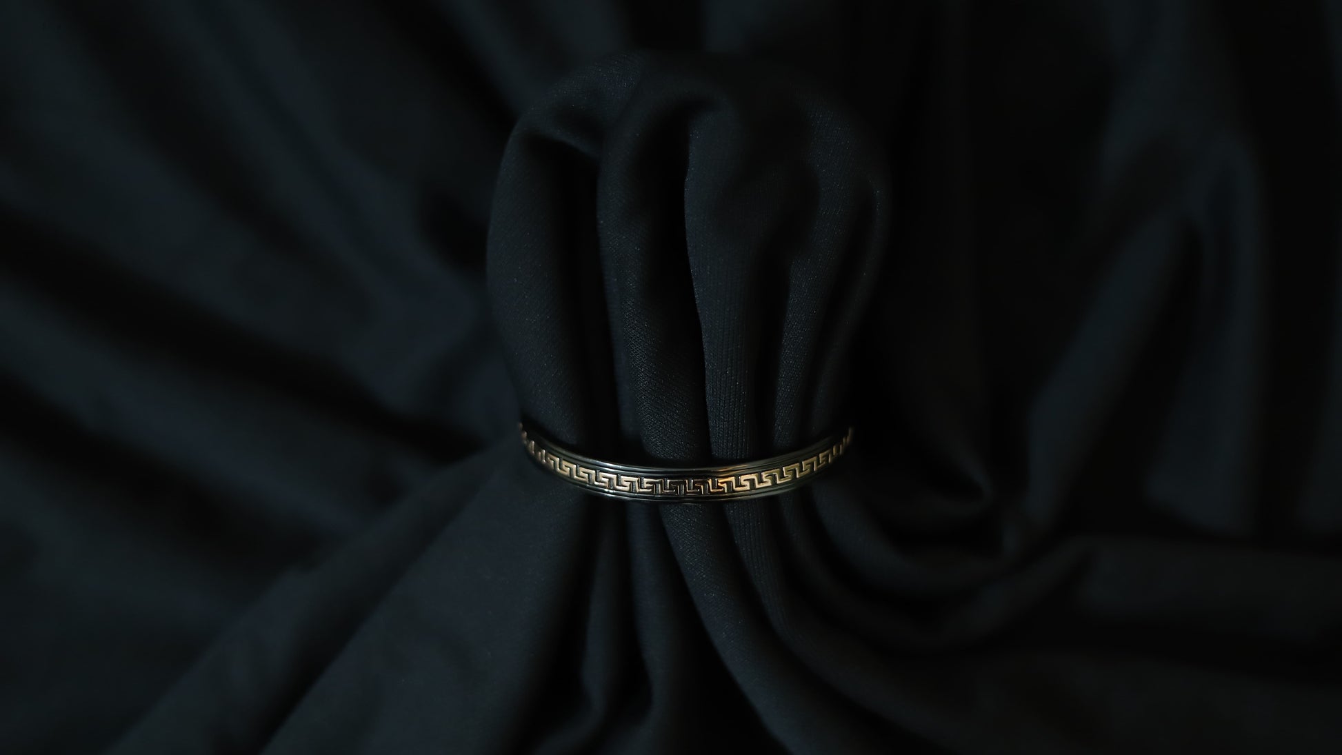 A black bangle bracelet with a gold Greek key pattern, wrapped around a dark black fabric bundle.