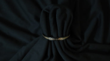A black bangle bracelet with a gold Greek key pattern, wrapped around a dark black fabric bundle.