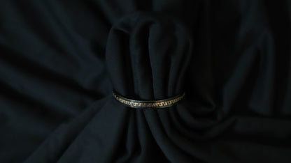 A black bangle bracelet with a gold Greek key pattern, wrapped around a dark black fabric bundle.