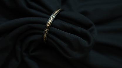 A slim metallic bangle bracelet with a Greek key pattern, resting on draped black fabric.