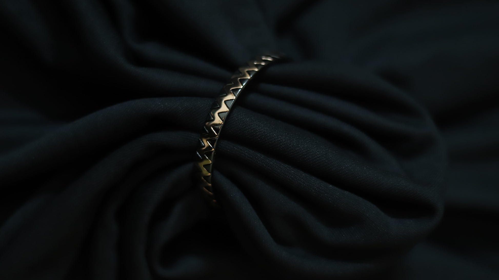 A close-up of a gold-toned metal chain bracelet with a repeating chevron or zigzag pattern, draped over dark black fabric.