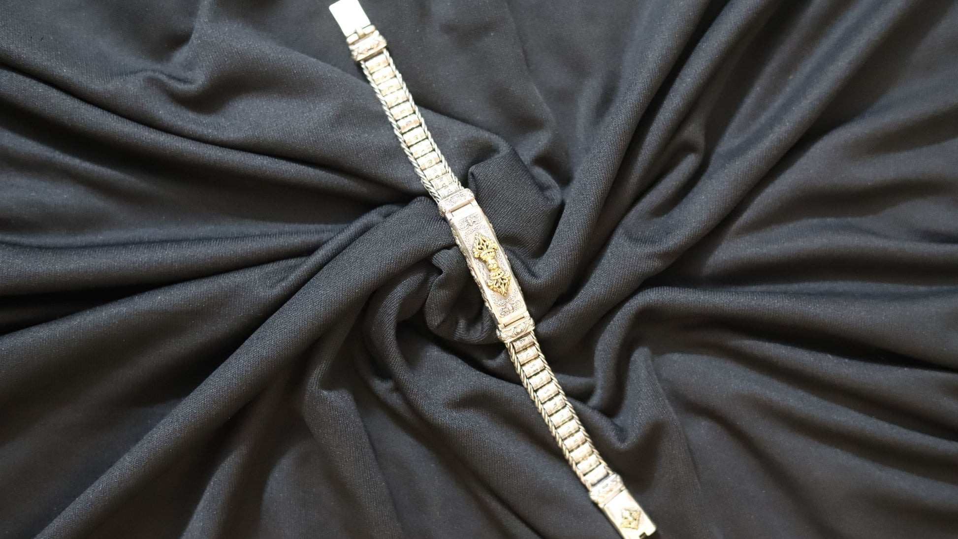 A silver-toned metal link bracelet with a central gold-toned decorative emblem, laid diagonally over draped black fabric.