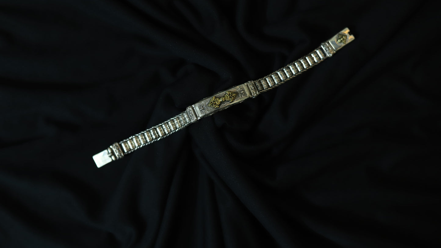 A silver-toned metal bracelet with a central gold-toned decorative plate, displayed diagonally on a draped black fabric background.
