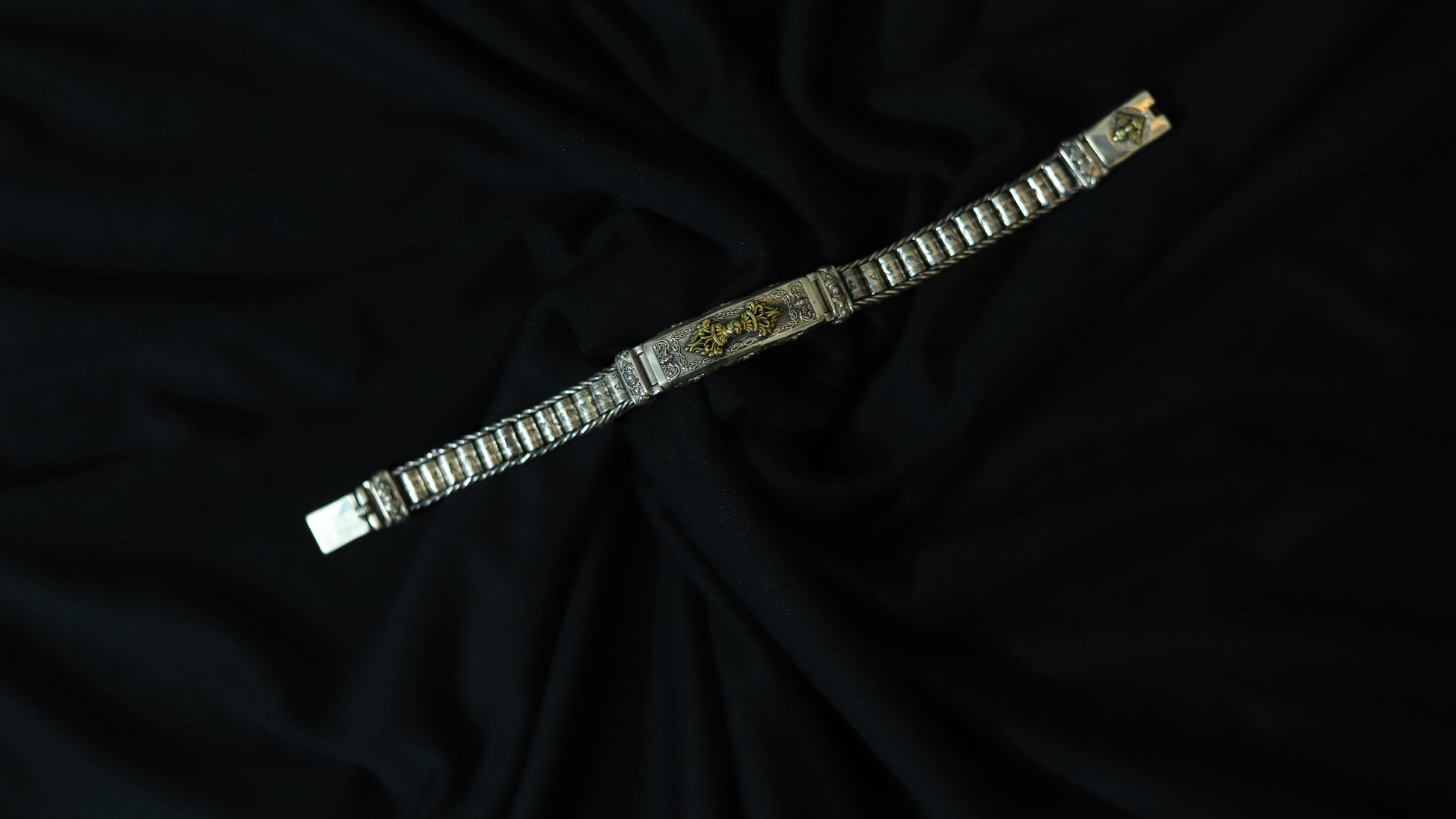 A silver-toned metal bracelet with a central gold-toned decorative plate, displayed diagonally on a draped black fabric background.