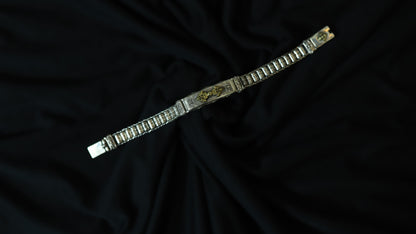 A silver-toned metal bracelet with a central gold-toned decorative plate, displayed diagonally on a draped black fabric background.