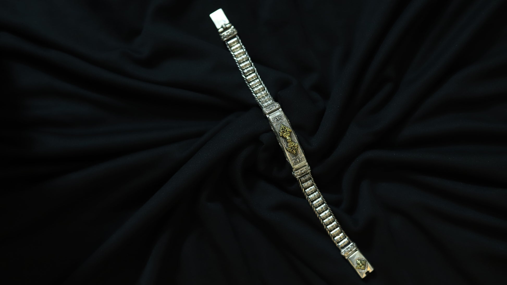 A silver-toned metal bracelet with textured rectangular links and gold-toned decorative details, displayed diagonally on a draped black fabric background.