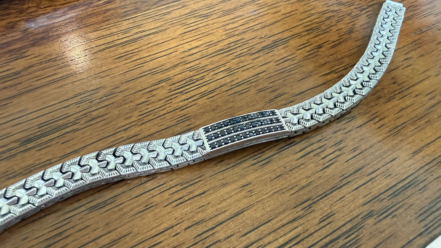 A silver-toned metal bracelet with a textured geometric pattern and a central black and silver inlay, displayed on a wooden surface.