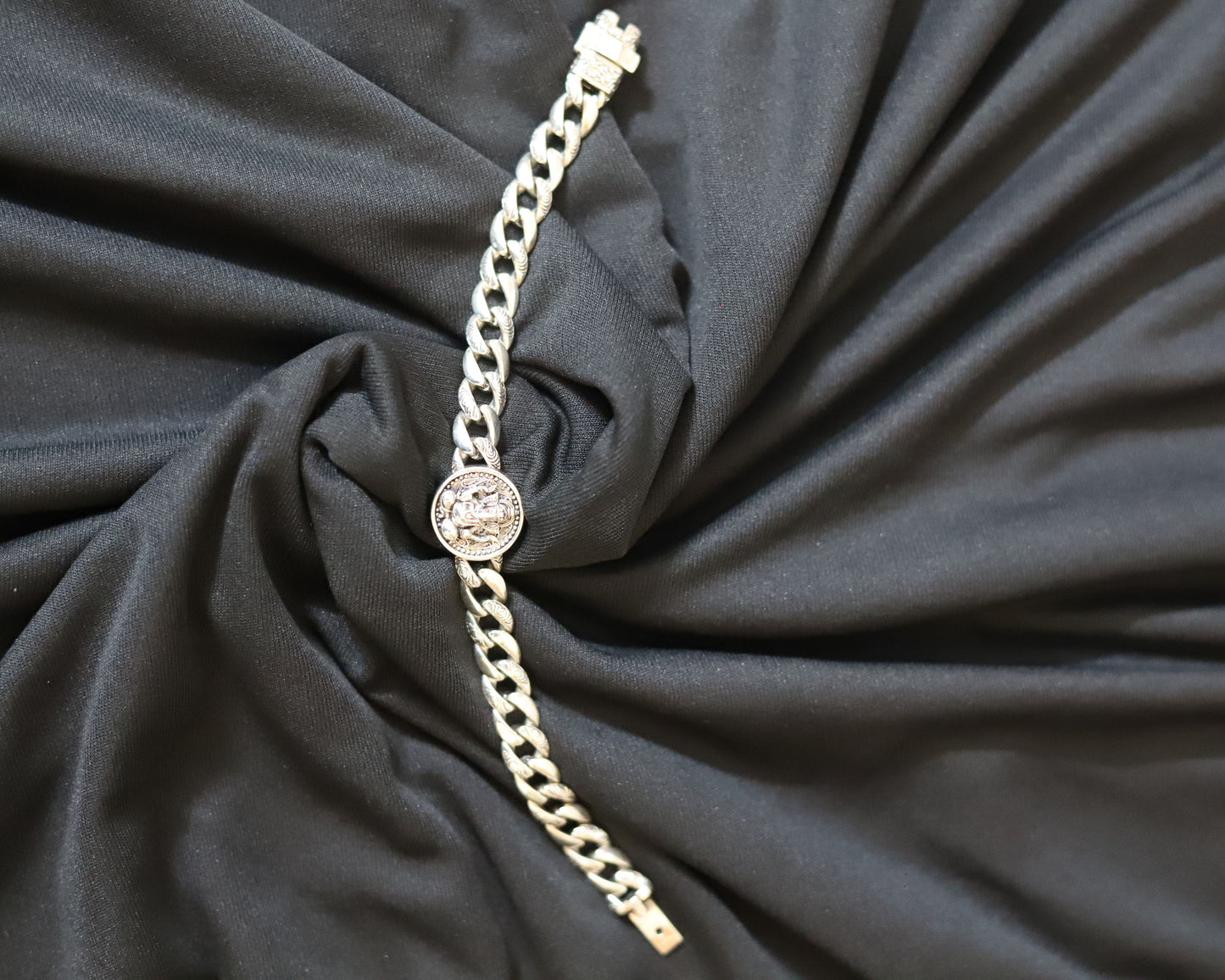 A silver-toned chain bracelet with a round bezel-set clear stone, displayed on a draped black fabric background.