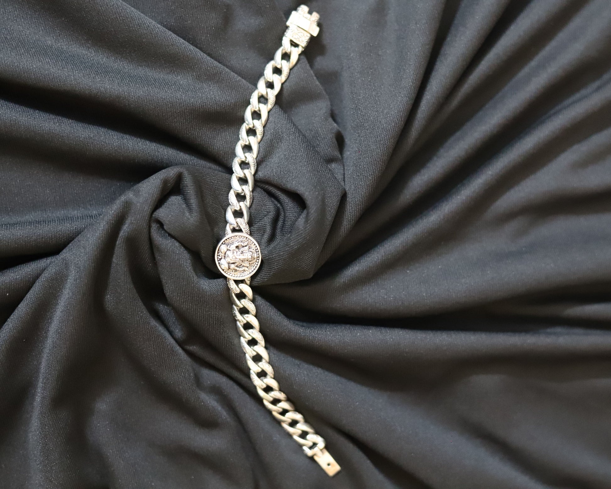 A silver-toned chain bracelet with a round bezel-set clear stone, displayed on a draped black fabric background.
