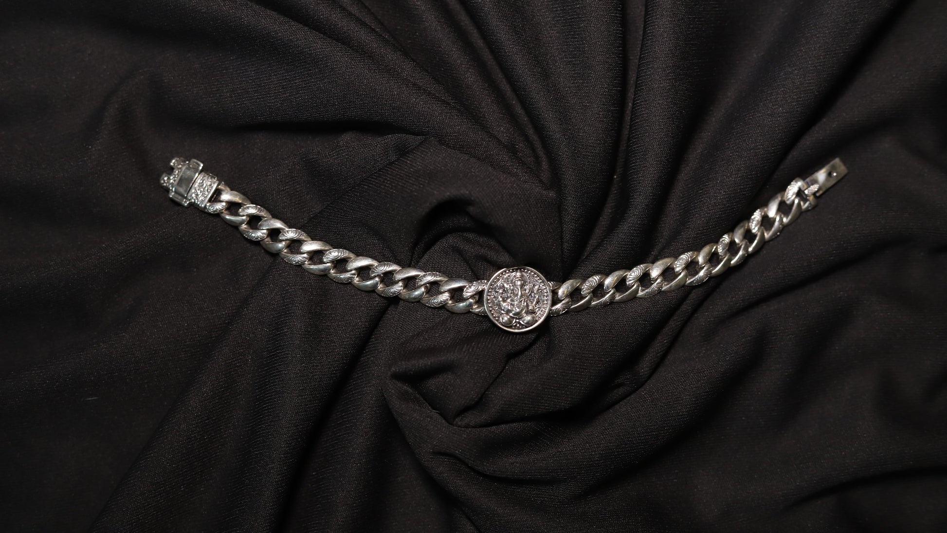 A silver-toned chain belt with a central round medallion and a clasp on the left, displayed on a draped black fabric background.