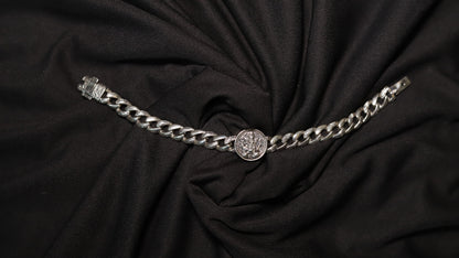 A silver-toned chain belt with a central round medallion and a clasp on the left, displayed on a draped black fabric background.