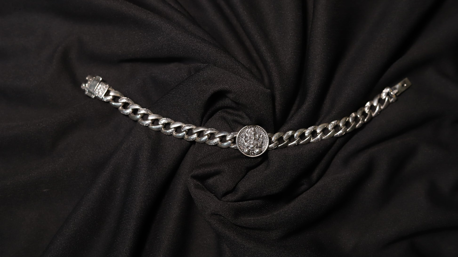 A silver chain belt with a central medallion and a buckle on the left, displayed on a draped black fabric background.